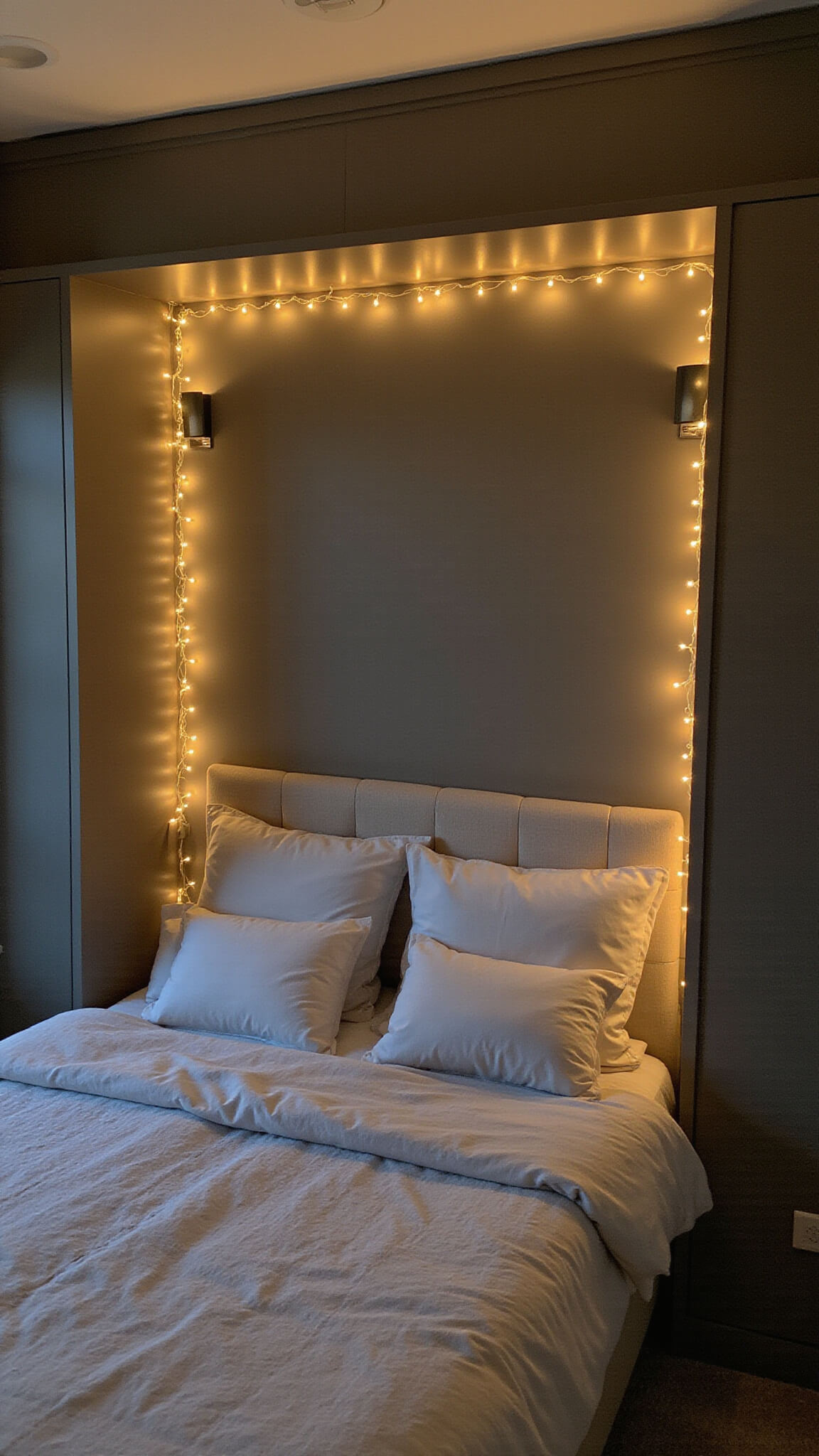 Cozy 8'x12' bedroom at twilight with warm layered lighting, string lights around tall bookshelf, sconces beside gray upholstered headboard, hidden LED strips, and neutral patterned textiles.