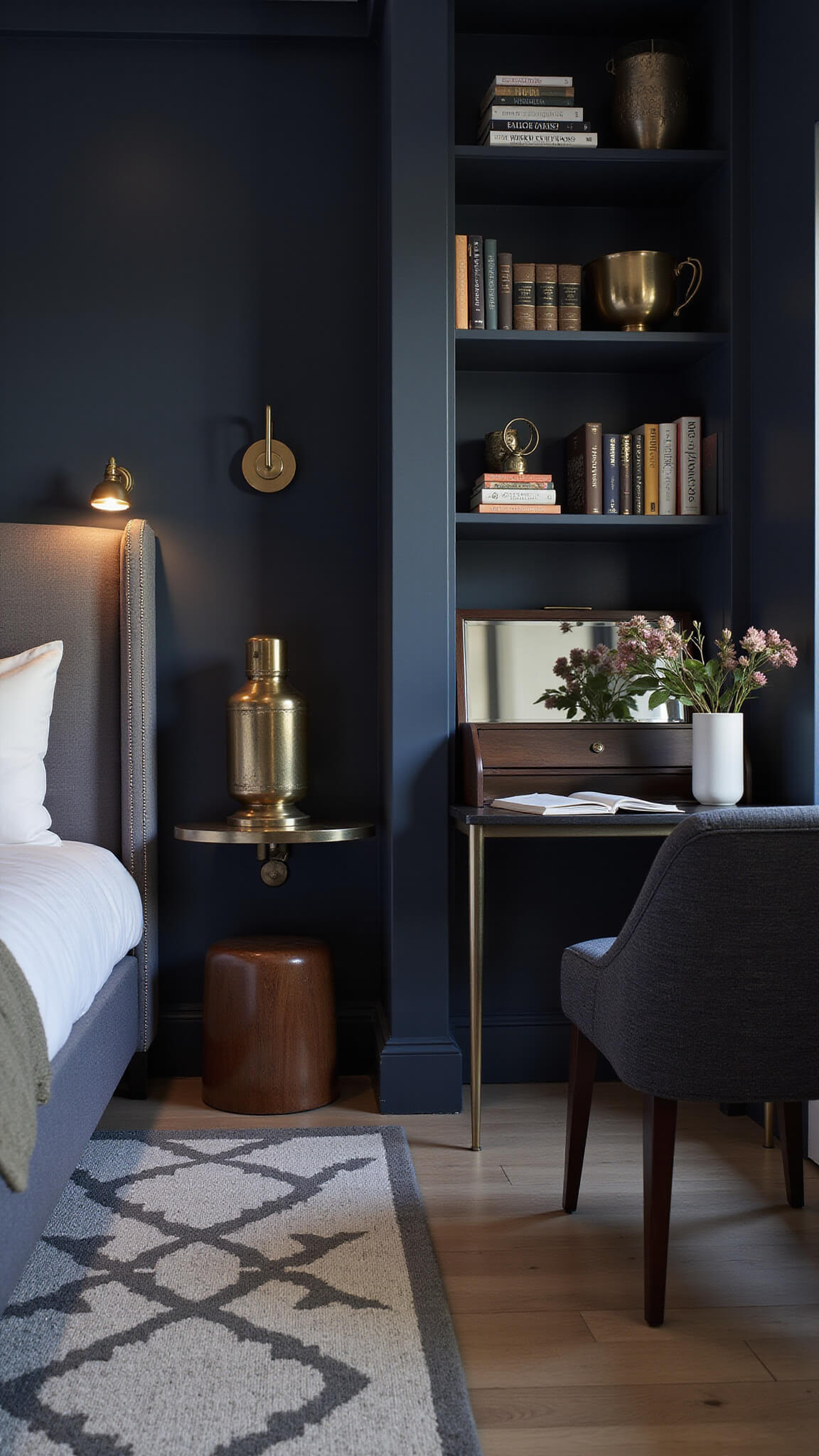 Contemporary guest suite at dusk with ambient lighting, low-profile queen bed against navy accent wall, warm brass sconces, grey and white geometric rug, floating shelves with brass decor, and mirror-backed secretary desk.