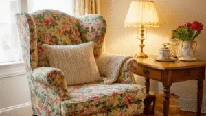 "A cozy corner with granny chic interior including a vintage floral wingback chair, a knitted afghan, a brass lamp on an oak table, all bathed in warm golden light."